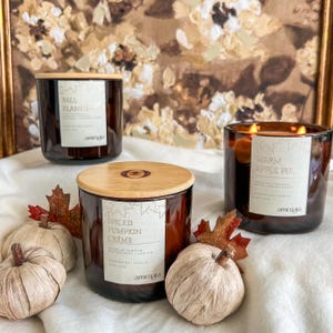 May include: Three scented candles in amber glass jars with wooden lids. The labels read "Fall Flannel", "Spiced Pumpkin Creme", and "Warm Apple Pie". Decorative pumpkins and fall leaves surround the candles, creating a cozy autumn theme.