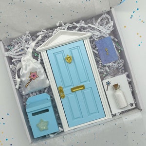 May include: A blue fairy door with a gold door handle and a white frame, a blue mailbox with a star, a white bag with a pink wand and star, a blue card with a gold key, and a small bottle with white glitter and a wand.