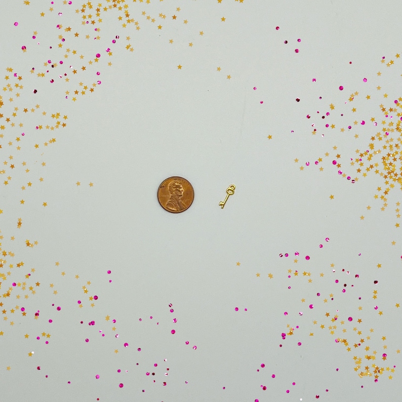 Peut inclure: Un petit charme de cl&eacute; en or sur un fond blanc avec des paillettes dor&eacute;es et roses. La cl&eacute; est &agrave; peu pr&egrave;s de la taille d'une pi&egrave;ce d'un centime d'euro.