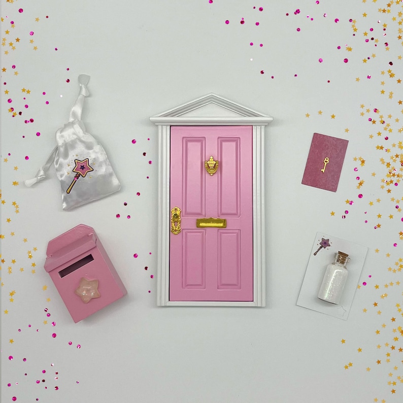 Peut inclure: Une porte de f&eacute;e rose avec une poign&eacute;e de porte dor&eacute;e et une bo&icirc;te aux lettres dor&eacute;e. Il y a un sac blanc avec un motif d'&eacute;toile et de baguette, une bouteille de paillettes et une carte avec une cl&eacute; dessus.