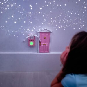 Peut inclure: Une petite porte rose avec une poign&eacute;e de porte dor&eacute;e et un cadre blanc est sur un mur blanc. Un enfant regarde la porte avec une expression r&ecirc;veuse. Le mur est d&eacute;cor&eacute; d'un effet de paillettes blanches scintillantes.
