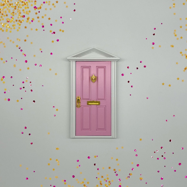 Peut inclure: Une porte miniature rose avec des accents dor&eacute;s et un cadre blanc sur un fond gris clair avec des paillettes dor&eacute;es et roses &eacute;parpill&eacute;es autour.