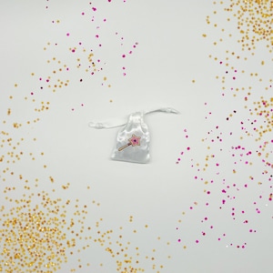 Peut inclure: Un sac en satin blanc avec cordon de serrage, d&eacute;cor&eacute; d'une baguette magique rose et blanche en forme d'&eacute;toile. Le sac est entour&eacute; de paillettes dor&eacute;es et roses.