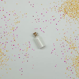 Peut inclure: Un petit flacon en verre avec un bouchon en li&egrave;ge rempli de paillettes blanches. Le flacon est entour&eacute; d'&eacute;toiles paillet&eacute;es dor&eacute;es et roses.