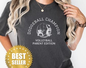 May include: A dark grey t-shirt with the text "Dodgeball Champion Volleyball Parent Edition" and a graphic of two people playing volleyball. The shirt has a gold star sticker that says "Best Seller".