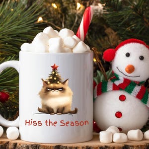 May include: White ceramic mug filled with marshmallows, a candy cane, and a grumpy cat wearing a Christmas tree hat. The mug has the text "Hiss the Season" in red. A snowman figure is next to the mug.