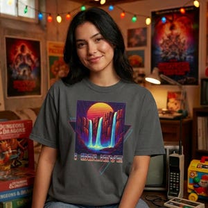 Könnte beinhalten: Graues T-Shirt mit Retro-Design, das einen Sonnenuntergang, Wasserfälle und den Text "I BELIEVE" zeigt. Das Design ist von einer geometrischen Form umrahmt. Das T-Shirt wird von einer Person getragen.