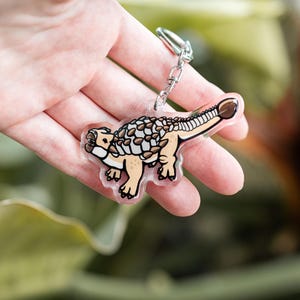 K&ouml;nnte beinhalten: Ein klarer Acryl-Schl&uuml;sselanh&auml;nger mit einem Cartoon-Ankylosaurus-Dinosaurier. Der Dinosaurier hat einen beigefarbenen K&ouml;rper, einen braunen Keulenschwanz und einen gemusterten R&uuml;cken. Der Schl&uuml;sselanh&auml;nger ist an einer silbernen Metallkette und einem Ring befestigt.
