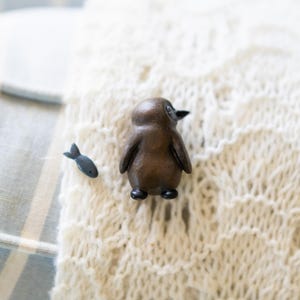 May include: A wooden penguin figurine with a black beak and black feet, sitting on a white knitted surface. A small black fish figurine is to the left of the penguin.