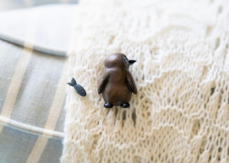 May include: A wooden penguin figurine with a black beak and black feet, sitting on a white knitted surface. A small black fish figurine is to the left of the penguin.