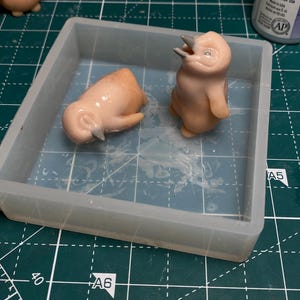 May include: Two small, light pink, ceramic penguin figurines in a clear silicone mold. The penguins are in different poses, one is standing upright with its mouth open and the other is lying on its side with its head down.