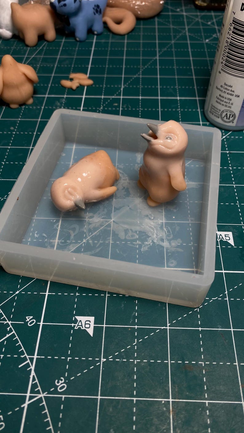 May include: Two small, light pink, ceramic penguin figurines in a clear silicone mold. The penguins are in different poses, one is standing upright with its mouth open and the other is lying on its side with its head down.