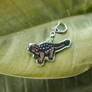 K&ouml;nnte beinhalten: Ein klarer Acryl-Schl&uuml;sselanh&auml;nger mit einem Ankylosaurus-Dinosaurier im Cartoon-Stil. Der Dinosaurier hat einen braunen K&ouml;rper, einen schwarz-wei&szlig; gemusterten R&uuml;cken und einen silbernen Schl&uuml;sselring. Der Schl&uuml;sselanh&auml;nger liegt auf einem gr&uuml;nen Blatt.