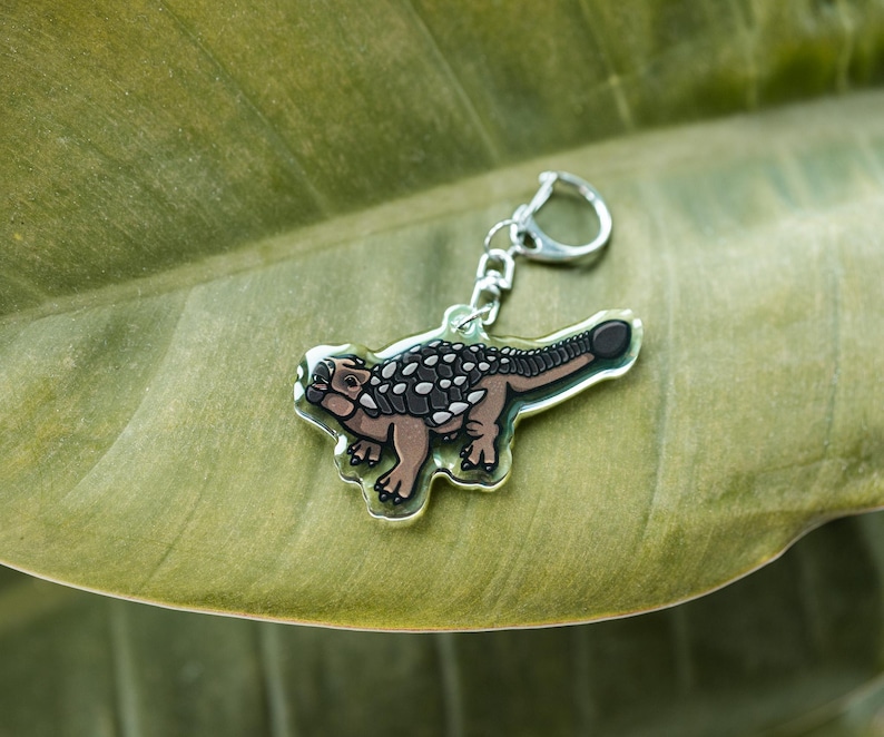 K&ouml;nnte beinhalten: Ein klarer Acryl-Schl&uuml;sselanh&auml;nger mit einem Ankylosaurus-Dinosaurier im Cartoon-Stil. Der Dinosaurier hat einen braunen K&ouml;rper, einen schwarz-wei&szlig; gemusterten R&uuml;cken und einen silbernen Schl&uuml;sselring. Der Schl&uuml;sselanh&auml;nger liegt auf einem gr&uuml;nen Blatt.