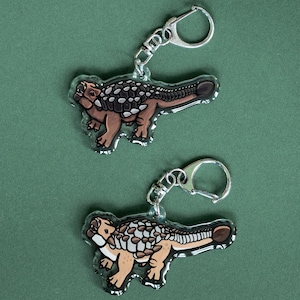 K&ouml;nnte beinhalten: Zwei transparente Acryl-Schl&uuml;sselanh&auml;nger mit einem Cartoon-Ankylosaurus-Dinosaurier-Design. Die Dinosaurier sind braun, beige und wei&szlig; mit einem dunkelbraunen Schwanz und R&uuml;cken. Jeder Schl&uuml;sselanh&auml;nger hat einen silberfarbenen Metall-Schl&uuml;sselring.