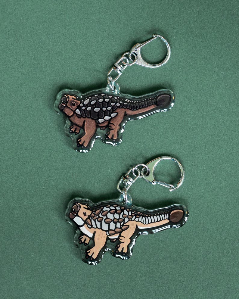 K&ouml;nnte beinhalten: Zwei transparente Acryl-Schl&uuml;sselanh&auml;nger mit einem Cartoon-Ankylosaurus-Dinosaurier-Design. Die Dinosaurier sind braun, beige und wei&szlig; mit einem dunkelbraunen Schwanz und R&uuml;cken. Jeder Schl&uuml;sselanh&auml;nger hat einen silberfarbenen Metall-Schl&uuml;sselring.