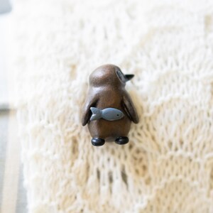 May include: A small wooden penguin figurine with a gray fish in its beak. The figurine is brown and has a white belly. It is sitting on a white knitted surface.