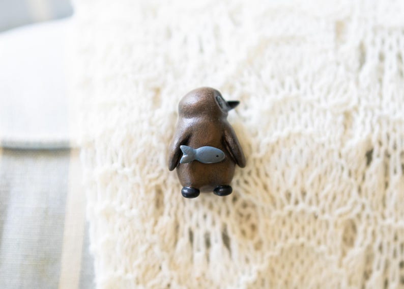 May include: A small wooden penguin figurine with a gray fish in its beak. The figurine is brown and has a white belly. It is sitting on a white knitted surface.