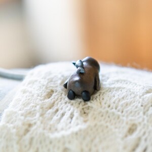 May include: A small, wooden figurine of a moose lying on a white knitted blanket. The moose is brown with black hooves and a silver nose.