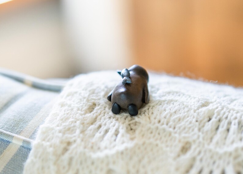 May include: A small, wooden figurine of a moose lying on a white knitted blanket. The moose is brown with black hooves and a silver nose.