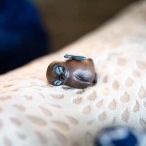 May include: A small, brown, ceramic figurine of a sleeping animal with blue eyes. The figurine is lying on a light brown, patterned surface.
