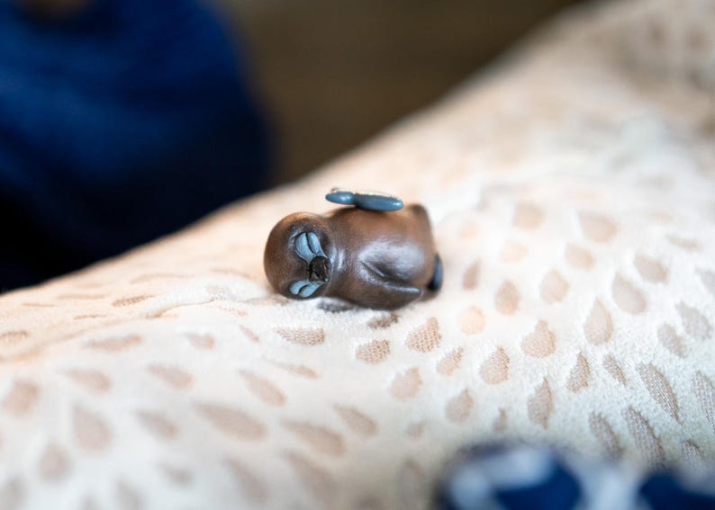 May include: A small, brown, ceramic figurine of a sleeping animal with blue eyes. The figurine is lying on a light brown, patterned surface.