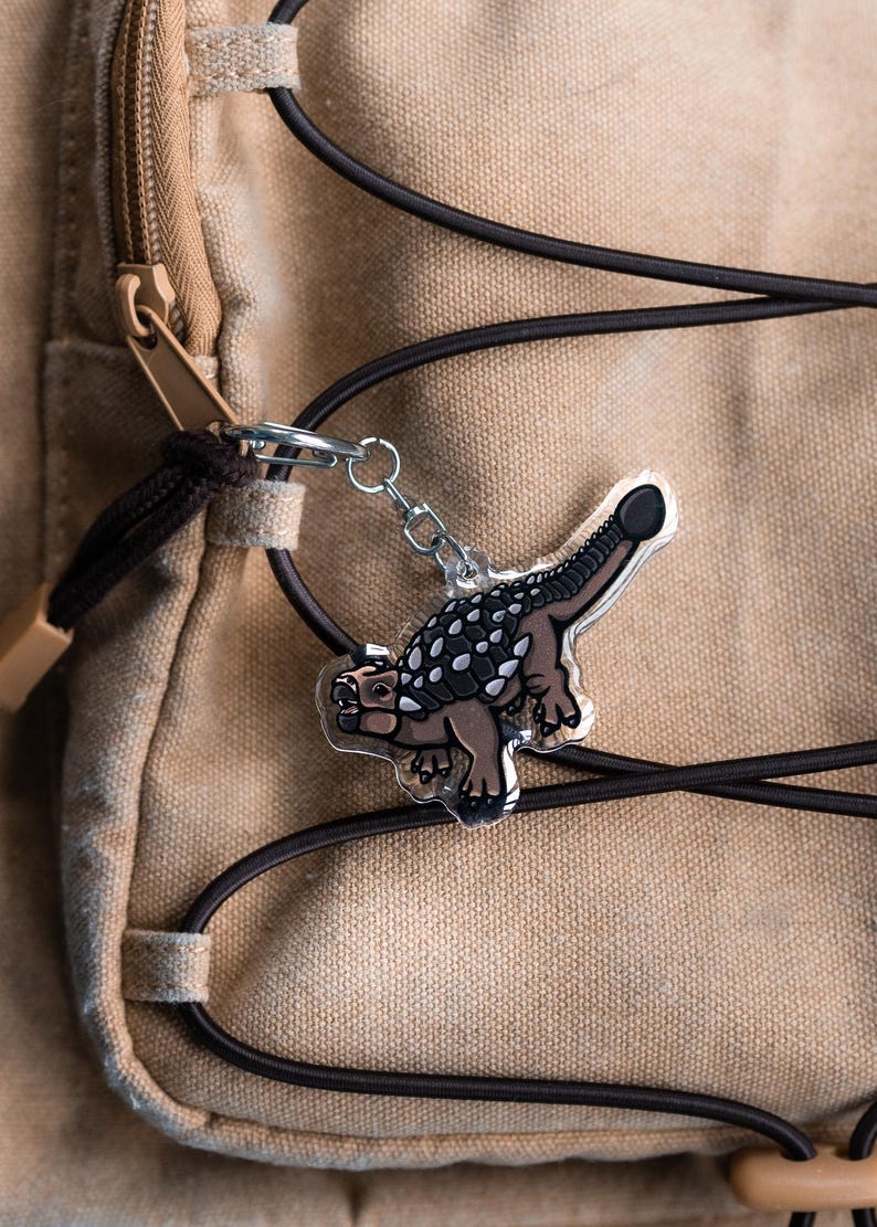 K&ouml;nnte beinhalten: Ein klarer Acryl-Schl&uuml;sselanh&auml;nger mit einem braunen und grauen Ankylosaurus-Dinosaurier-Design. Der Schl&uuml;sselanh&auml;nger ist an einer silbernen Kette und einem Metallverschluss befestigt. Der Hintergrund ist ein beigefarbener Canvas-Rucksack mit schwarzen Kordeln.