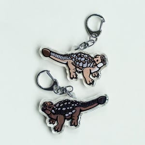 K&ouml;nnte beinhalten: Zwei transparente Acryl-Schl&uuml;sselanh&auml;nger mit einem Cartoon-Ankylosaurus-Dinosaurier-Design. Die Dinosaurier sind in Braun-, Beige- und Wei&szlig;t&ouml;nen gehalten und haben einen silbernen Schl&uuml;sselanh&auml;nger. Die Schl&uuml;sselanh&auml;nger befinden sich auf wei&szlig;em Hintergrund.
