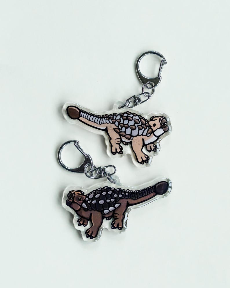 K&ouml;nnte beinhalten: Zwei transparente Acryl-Schl&uuml;sselanh&auml;nger mit einem Cartoon-Ankylosaurus-Dinosaurier-Design. Die Dinosaurier sind in Braun-, Beige- und Wei&szlig;t&ouml;nen gehalten und haben einen silbernen Schl&uuml;sselanh&auml;nger. Die Schl&uuml;sselanh&auml;nger befinden sich auf wei&szlig;em Hintergrund.