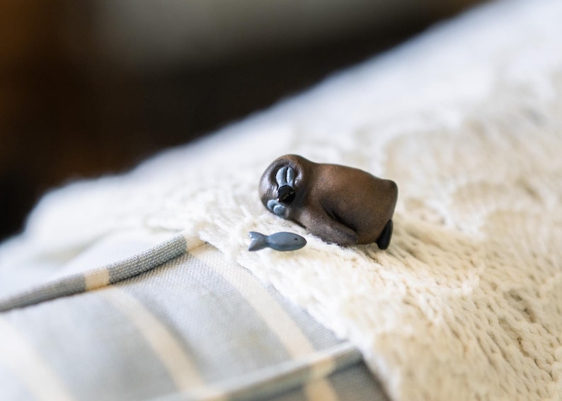 May include: Close-up view of a small, dark brown figurine resembling a sleeping animal, possibly a sea otter or similar creature, with a tiny, light blue fish figurine beside it. Both figurines appear to be crafted from a smooth material, possibly resin or ceramic. The animal figurine is positioned on a light beige, textured fabric surface with subtle stripes of light blue. The overall style suggests a miniature collectible or decorative item with a whimsical, nature-inspired theme.