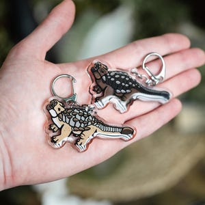 K&ouml;nnte beinhalten: Zwei transparente Acryl-Schl&uuml;sselanh&auml;nger mit einer Cartoon-Illustration eines Ankylosaurus-Dinosauriers. Die Dinosaurier sind beige, braun und schwarz mit silbernen Schl&uuml;sselringen. Die Schl&uuml;sselanh&auml;nger werden in einer Hand gehalten.