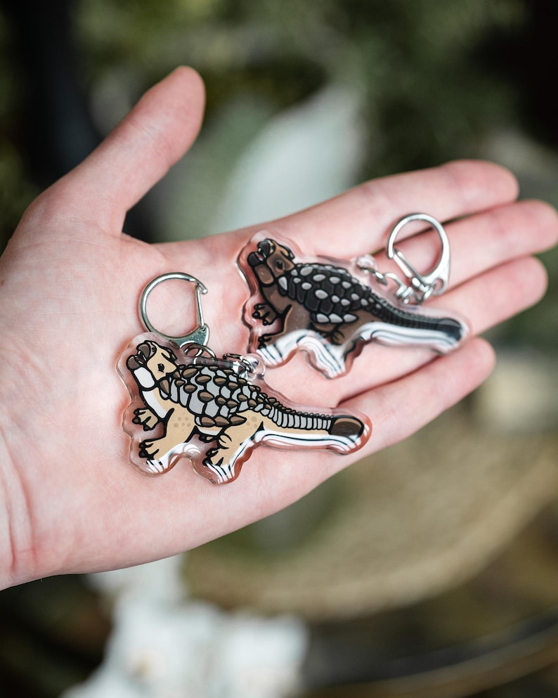 K&ouml;nnte beinhalten: Zwei transparente Acryl-Schl&uuml;sselanh&auml;nger mit einer Cartoon-Illustration eines Ankylosaurus-Dinosauriers. Die Dinosaurier sind beige, braun und schwarz mit silbernen Schl&uuml;sselringen. Die Schl&uuml;sselanh&auml;nger werden in einer Hand gehalten.