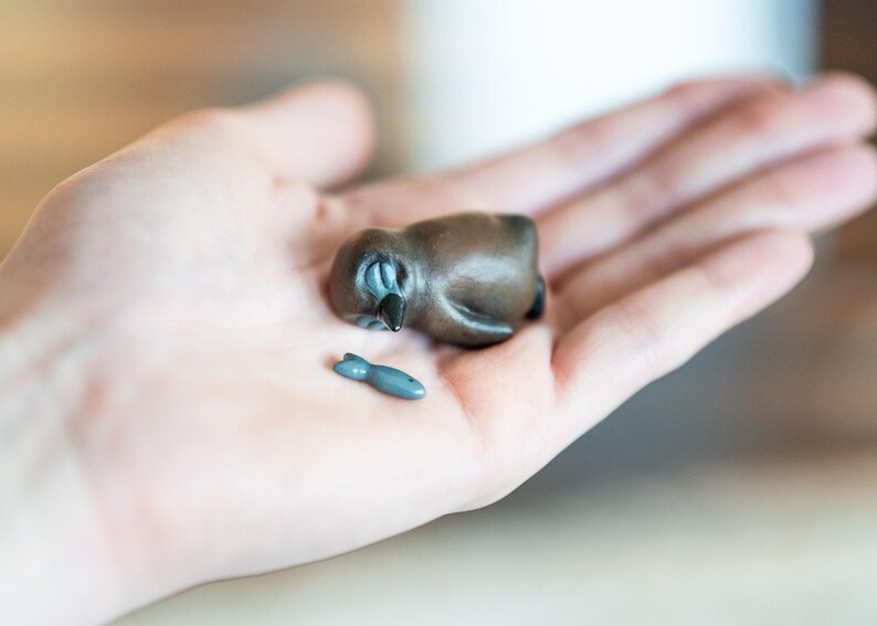 May include: A small, brown ceramic penguin figurine with closed eyes, lying on its side. A small, gray ceramic fish figurine is next to the penguin.
