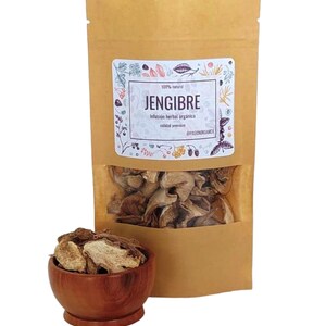 May include: A brown paper bag with a white label that says "JENGIBRE" and "Infusion herbal organica calidad premium" in Spanish. The bag contains dried ginger pieces. A small wooden bowl filled with dried ginger pieces is in the foreground.