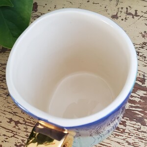 K&ouml;nnte beinhalten: Eine wei&szlig;e Keramiktasse mit goldenem Henkel und blau-t&uuml;rkisem Design. Die Innenseite der Tasse ist wei&szlig;. Die Tasse steht auf einer verwitterten, hellen Holzoberfl&auml;che.