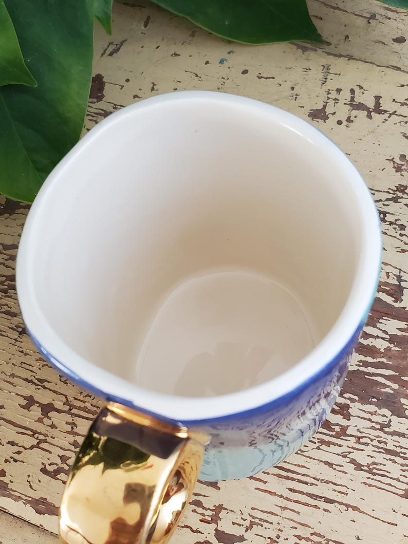 K&ouml;nnte beinhalten: Eine wei&szlig;e Keramiktasse mit goldenem Henkel und blau-t&uuml;rkisem Design. Die Innenseite der Tasse ist wei&szlig;. Die Tasse steht auf einer verwitterten, hellen Holzoberfl&auml;che.