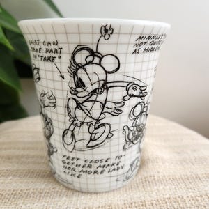 Op de afbeelding: Witte keramische beker met een zwarte schets van Minnie Mouse en handgeschreven notities. De beker heeft rasterlijnen en de tekst "Shirt can take part in 'take'" en "Feet close together make her more lady like."
