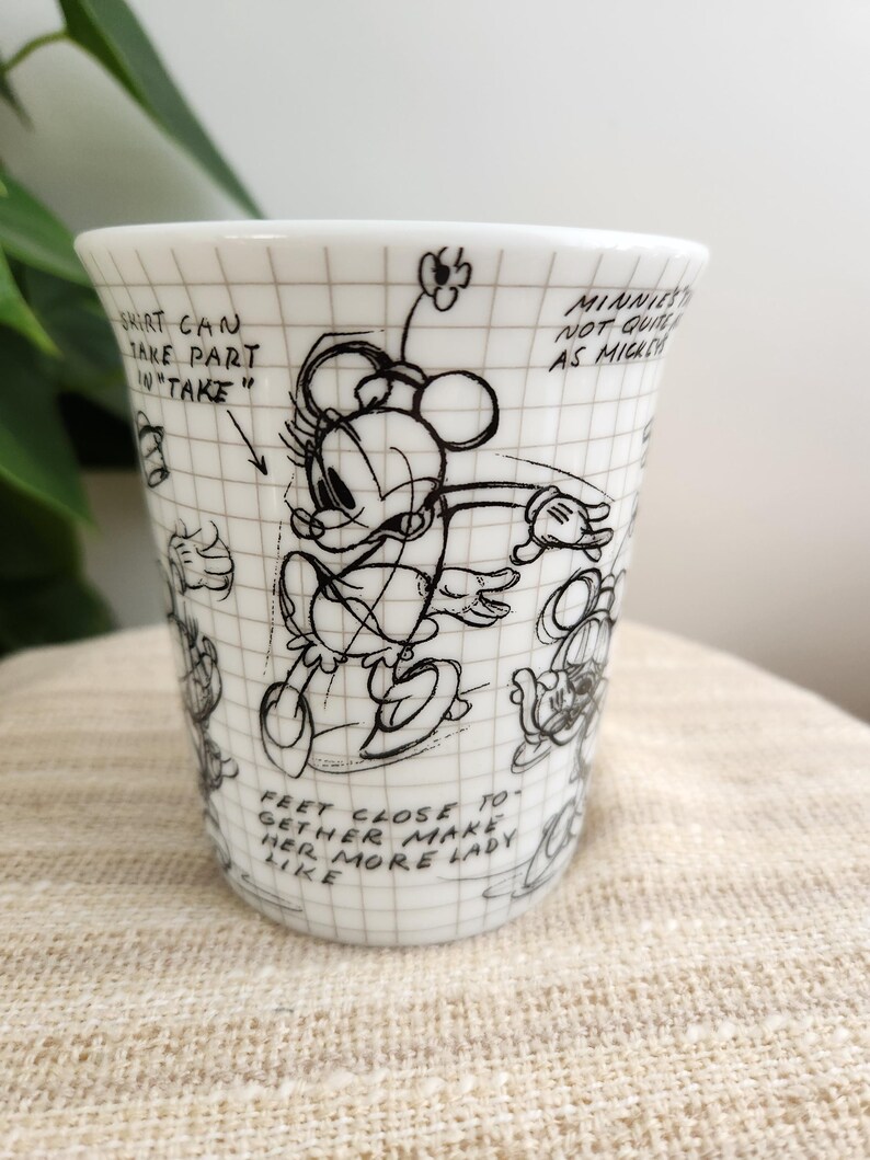 Op de afbeelding: Witte keramische beker met een zwarte schets van Minnie Mouse en handgeschreven notities. De beker heeft rasterlijnen en de tekst "Shirt can take part in 'take'" en "Feet close together make her more lady like."