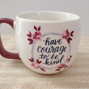 Op de afbeelding: Witte keramische mok met een bordeauxrode handgreep. De mok heeft een bloemenmotief met roze en blauwe accenten. De tekst "have courage to be kind" is op de mok gedrukt.