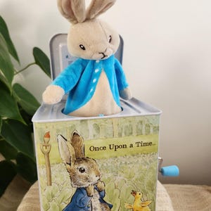 May include: A Peter Rabbit themed Jack-in-the-box toy. The box is decorated with illustrations of Peter Rabbit and a duckling. The plush rabbit wears a blue jacket. The box has a blue crank and the text "Once Upon a Time..."