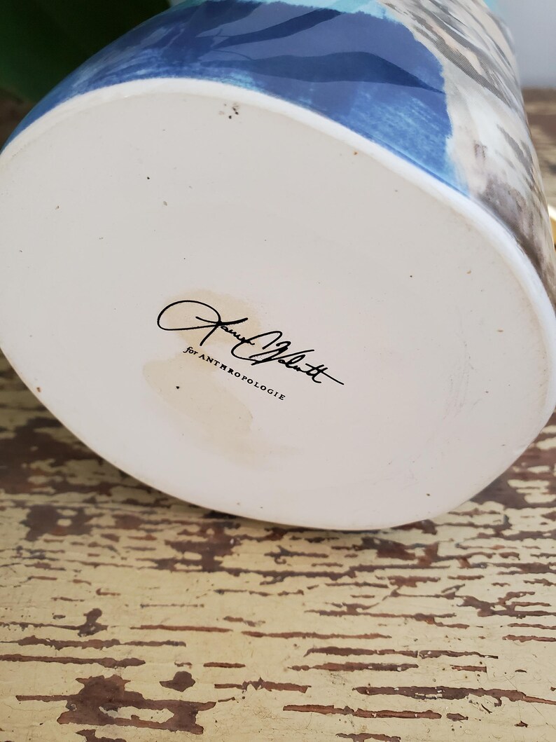 K&ouml;nnte beinhalten: Eine Keramikvase mit wei&szlig;em Boden und blau-grauem Anstrich. Der Boden der Vase tr&auml;gt eine Signatur und den Text "For ANTHROPOLOGIE". Die Vase steht auf einer verwitterten, gelb lackierten Holzoberfl&auml;che.