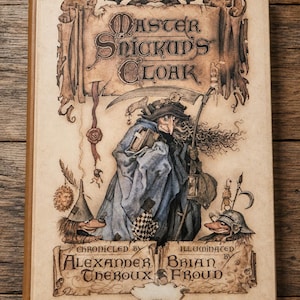 May include: A hardcover book titled "Master Snickup's Cloak" with illustrations. The cover features a robed figure with a scythe, a lantern, and other whimsical characters. The book is "chronicled by Alexander Theroux" and "illuminated by Brian Froud."