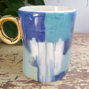 K&ouml;nnte beinhalten: Eine Keramiktasse mit goldfarbenem Henkel und wei&szlig;em Innenraum. Die Au&szlig;enseite weist ein abstraktes Design in Blau, T&uuml;rkis und Wei&szlig; auf. Die Tasse steht auf einer verwitterten Holzoberfl&auml;che.