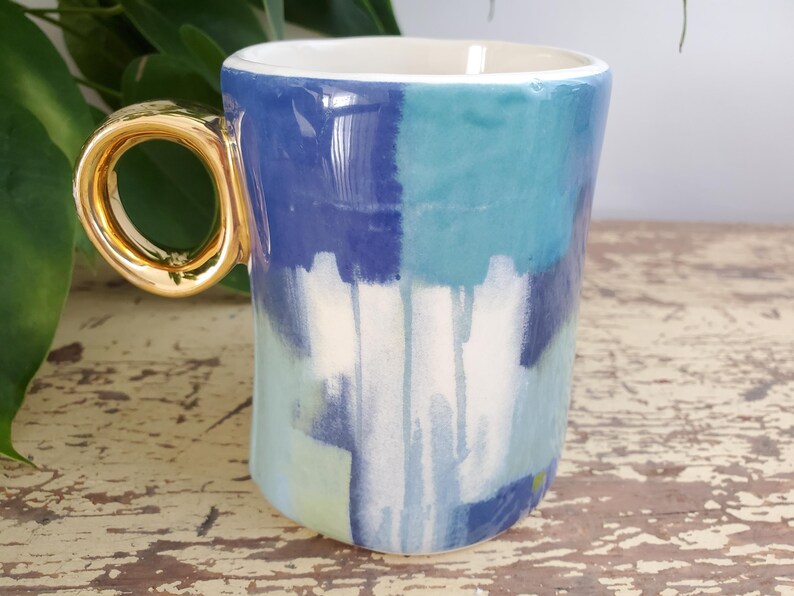 K&ouml;nnte beinhalten: Eine Keramiktasse mit goldfarbenem Henkel und wei&szlig;em Innenraum. Die Au&szlig;enseite weist ein abstraktes Design in Blau, T&uuml;rkis und Wei&szlig; auf. Die Tasse steht auf einer verwitterten Holzoberfl&auml;che.