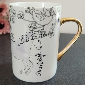May include: A white ceramic mug featuring a black line art illustration of a unicorn surrounded by floral branches. The word Magical is printed in black script below the unicorn. The mug has a shiny gold-colored handle and a smooth finish.