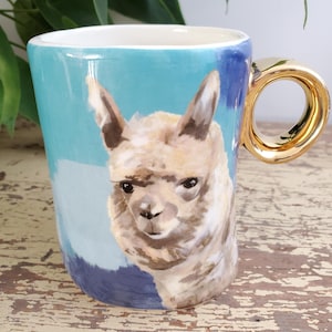 K&ouml;nnte beinhalten: Eine Keramik-Tasse mit goldenem Henkel und einem gemalten Bild eines Alpakas. Die Tasse hat einen hellblauen und dunkelblauen Hintergrund. Das Alpaka ist in Braun- und Wei&szlig;t&ouml;nen gemalt.