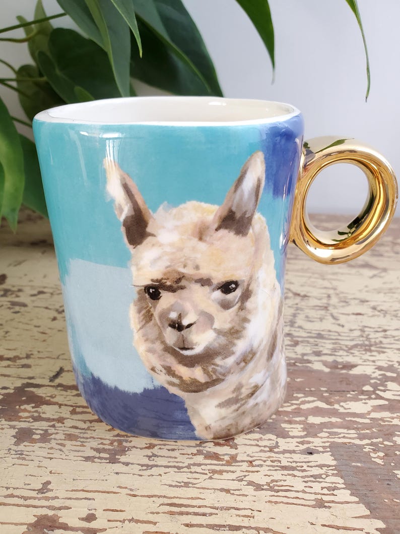 K&ouml;nnte beinhalten: Eine Keramik-Tasse mit goldenem Henkel und einem gemalten Bild eines Alpakas. Die Tasse hat einen hellblauen und dunkelblauen Hintergrund. Das Alpaka ist in Braun- und Wei&szlig;t&ouml;nen gemalt.