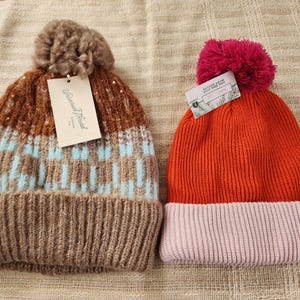 May include: Two knit beanies. One is brown, light blue, and tan with a brown pom-pom. The other is orange with a pink brim and a pink pom-pom. Both have tags attached.