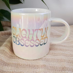 Puede incluir: Taza de cerámica blanca iridiscente con el texto "SLIGHTLY OBSESSED" en letras coloridas de estilo retro. La taza tiene un asa curva y un acabado nacarado, que refleja un arcoíris de colores. Una taza de café elegante y divertida.
