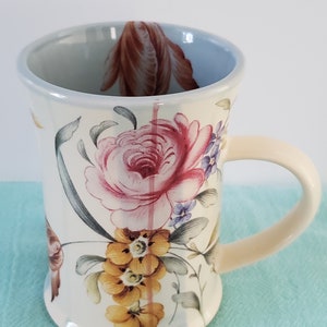 May include: A white ceramic mug with a floral design featuring pink, yellow, and blue flowers. The mug has a cream-colored handle.